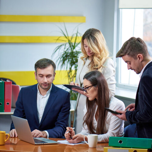 A group of two women and two men sit around a table and work on a laptop